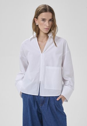 Žena s rovnými svetlohnedými vlasmi, ktorá má na sebe veľké strieborné náušnice, voľnú bielu košeľu s gombíkmi a náprsným vreckom a modré rifle s vysokým pásom.