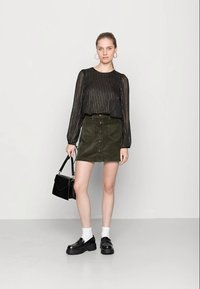 Blusa preta riscada com mangas compridas, saia de veludo cotelê verde-azeitona com botões frontais, meias brancas, sapatos pretos robustos e uma bolsa preta.