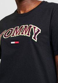 Camiseta de algodón negra con "TOMMY" en un bordado en negrita rojo y verde, con un pequeño emblema rojo, blanco y azul debajo. Manga corta.