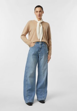 Donna che indossa un maglione beige, una sciarpa bianca al collo, jeans blu a gamba larga e scarpe nere, in piedi con una mano in tasca.
