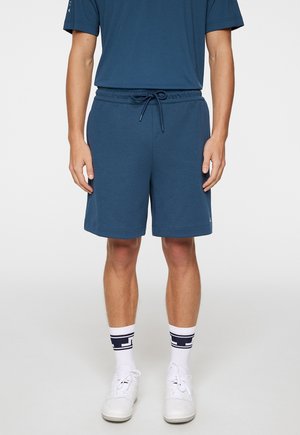 Herre blå bomulds shorts med elastisk talje og snøre. Har sidelommer og en afslappet pasform, parret med hvide sneakers og sokker.