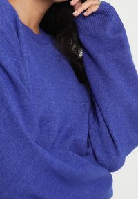 Personne portant un pull bleu en maille douce à manches longues, avec une main levée près du visage et des cheveux foncés partiellement visibles.