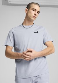 Tricou scurt cu mânecă scurtă de culoare gri deschis, cu guler tip crew și un logo Puma negru pe piept. Are un material neted și o croială lejeră.
