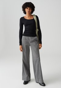 Vrouw met krullend haar, gekleed in een zwarte top met lange mouwen, grijze broek met wijde pijpen, zwarte schoenen en met een groene schoudertas, staat tegen een effen achtergrond.