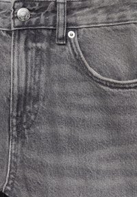 Primer plano de unos vaqueros de denim gris que muestran el bolsillo frontal, el lazo del cinturón, los remaches de metal, el botón y los detalles de la textura de la tela.