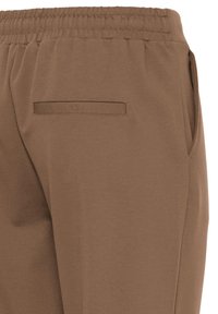Pantalon marron avec taille élastique, poche arrière passepoilée et poches latérales, vue rapprochée de la partie supérieure de la hanche et de la taille.
