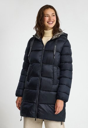 Donna sorridente che indossa un cappotto imbottito nero con cappuccio sopra un maglione beige a collo alto, in piedi davanti a uno sfondo bianco uniforme.