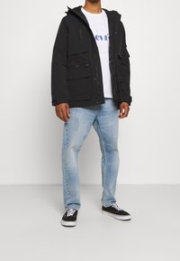 Černá kapucínová bunda s mnoha kapsami, zipem vpředu a nastavitelným manžetami, nošena přes bílé tričko a světle modré džíny. Černé tenisky.