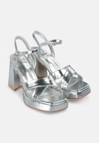Sandalias de tacón alto plateadas con punta abierta, diseño de tiras cruzadas y un robusto tacón cuadrado. Material metálico suave con un acabado brillante.