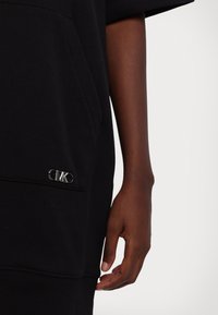 Gros plan d'une main à la peau foncée à côté d'une poche de vêtement noire avec un petit logo métallique "CK".