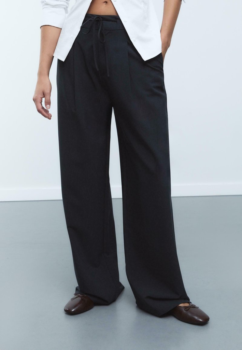 Pantalon noir à jambes larges avec un motif rayé vertical subtil, taille à cordon de serrage et plis, associé à des chaussures plates marron foncé.