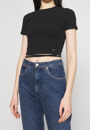 Femme portant un haut court à manches courtes noir avec des liens enveloppants et un jean bleu taille haute sur un fond uni.