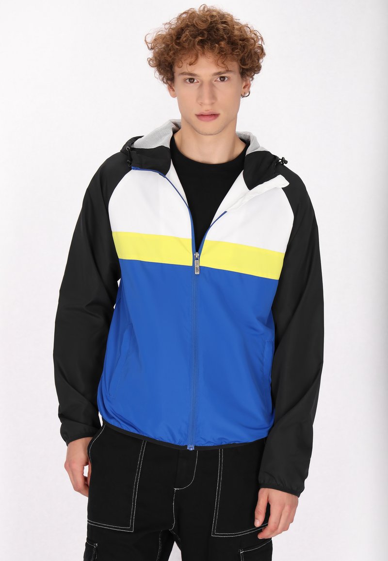 Giacca a vento colorata con corpo blu, strisce bianche e gialle e maniche nere. Presenta una chiusura con zip e un cappuccio regolabile.