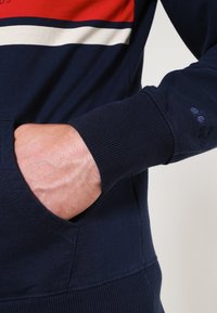 Sweatshirt bleu marine avec un design rayé rouge et crème sur la poitrine, des poignets côtelés et une poche kangourou avec une finition texturée.