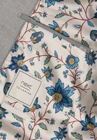 Musterstoff mit blauen und goldenen Blumen auf einem hellen Hintergrund, mit grau gefüttert und einem Etikett, das "next TAILORING" liest.