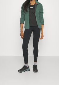 Giacca in pile verde con zip sopra una maglietta nera e leggings neri. Il modello indossa scarpe da ginnastica grigie e nere. Logo bianco sul petto e sulla gamba.