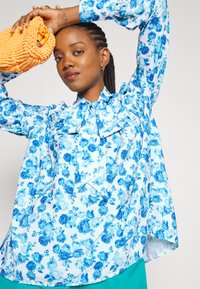 Blusa floral azul com um grande laço no colarinho, com mangas longas e um corte descontraído. Acompanhada de uma clutch texturizada laranja.