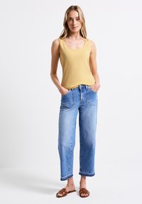 Top sin mangas amarillo, jeans de mezclilla anchos claros y sandalias planas marrones. Los jeans tienen bolsillos y detalles de dobladillo cosido.