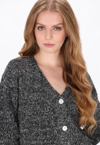 Šedý pletený cardigan s výstřihem do V, který má tři bílé knoflíky a texturovanou tkaninu. Dlouhé rukávy s volným střihem.