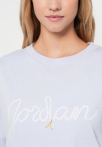 Close-up van een persoon die een licht lila sweatshirt draagt met het woord "Jordan" gespeld in witte pailletten en een klein gouden Jumpman-logo.