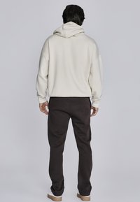 Világos bézs színű, laza szabású kapucnis pulóver, nagy kapucnival és bordás mandzsettákkal. Sötétbarna tréningnadrággal és világos lábbelivel párosítva.