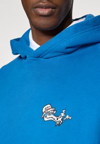 Μπλε φούτερ με σχέδιο γελοιογραφίας δράκου που φοράει καπέλο και έχει λεπτομέρειες καπνού. Μαλακό ύφασμα με χαλαρή εφαρμογή και ρυθμιζόμενη κουκούλα.