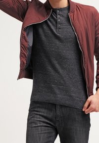 Burgundská bomber jacket na zip přes tmavě šedou henley košili, se zapínáním na knoflíky a žebrovanými manžetami, v kombinaci s tmavými džíny. Hladký povrch látky.