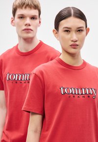Camisetas de algodón rojo con cuello redondo, con logo "tommy" bordado y texto "JEANS". Textura suave, ajuste relajado, costuras visibles.