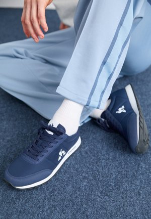 Zapatillas navy con una textura suave, detalles blancos y un logo en el lateral. Combinadas con pantalones de estilo deportivo azul claro y calcetines blancos.