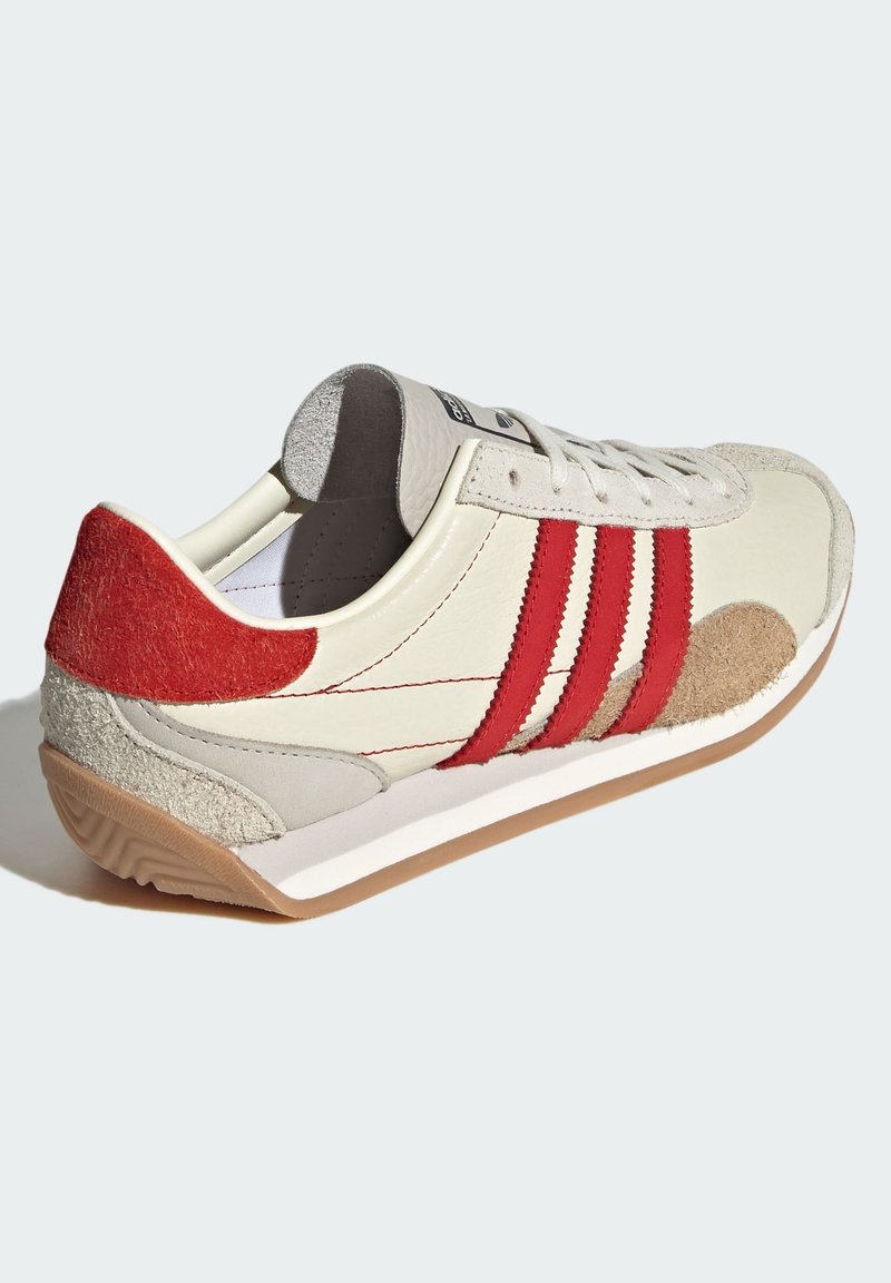 Zapatilla crema con rayas y detalles en rojo, talón de ante, suela de goma y detalles cosidos. Presenta una puntera redonda y un diseño con cordones.