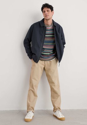 Chaqueta bomber navy sobre un suéter de rayas multicolores, combinada con pantalones beiges y zapatillas blancas con suelas de goma. De pie contra una pared.