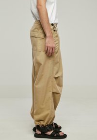 Beige cargo bukser har en elastisk talje med snor, sidelommer og elastiske manchetter, parret med sorte sandaler med stropper.