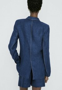 Syväsininen denim-takki, jossa on valkoiset ompeleet, takana yksi halkio ja pitkät hihat, yhdistettynä samasta kankaasta valmistettuihin shortseihin.