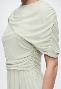 Κοντινή λήψη ενός ανοιχτό πράσινου φόρεματος με ρυτιδωμένο ύφασμα, φουσκωτό μανίκι και λεπτομέρειες με συγκεντρωμένο ύφασμα στο μπούστο και τη μέση.