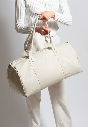 Borsa da viaggio in tela beige con doppie maniglie, cuciture rinforzate e una cerniera dorata. Presenta bretelle decorative e un design con base arrotondata.