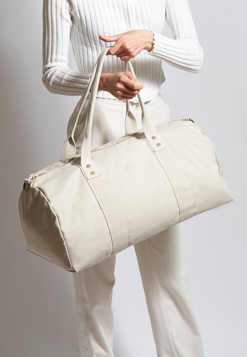 Beige canvas duffeltas met dubbele handvatten, versterkte stiksels en een gouden rits. Heeft accentriemen en een afgeronde bodemontwerp.