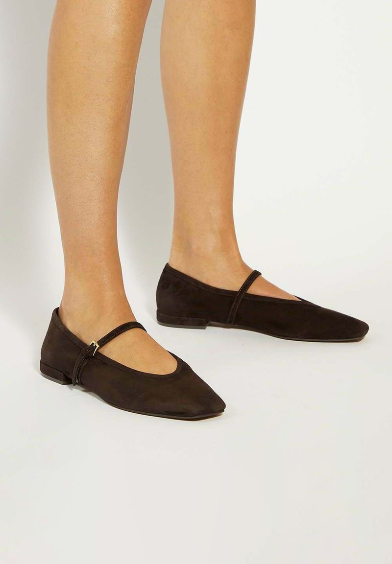 Benen in donkerbruin suède Mary Jane flats met een dunne band en een kleine gouden sluiting, staand op een lichtgekleurde vloer.