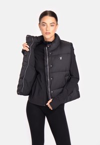 Črn puffer brezrokavnik z visokim ovratnikom, sprednjim zadrgo in zaponkami. Ima diamantno prešito vzorčenje in logo na levem prsnem delu.