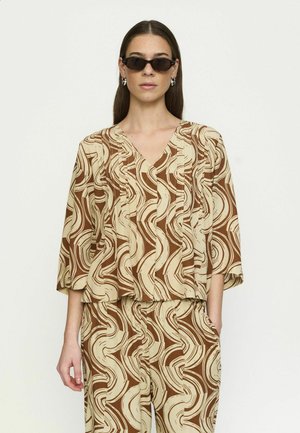 Naine, kes kannab pruuni ja kreemikat abstraktse keerisemustriga lahtist pluusi ja sobivaid pükse, mustade kassisilma päikeseprillidega ning hõbedaste rõngakujuliste kõrvarõngastega.