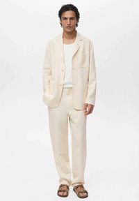 Lyse beige jakkesæt lavet af linned, med en enkeltknap blazer og brede bukser. Bæres med en hvid skjorte og brune sandaler.
