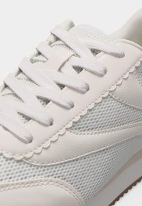 Λευκό αθλητικό παπούτσι με λεπτομέρειες από δίχτυ και δέρμα, περίτεχνη τρέσα και επίπεδα κορδόνια δεμένα, σε λευκό φόντο.