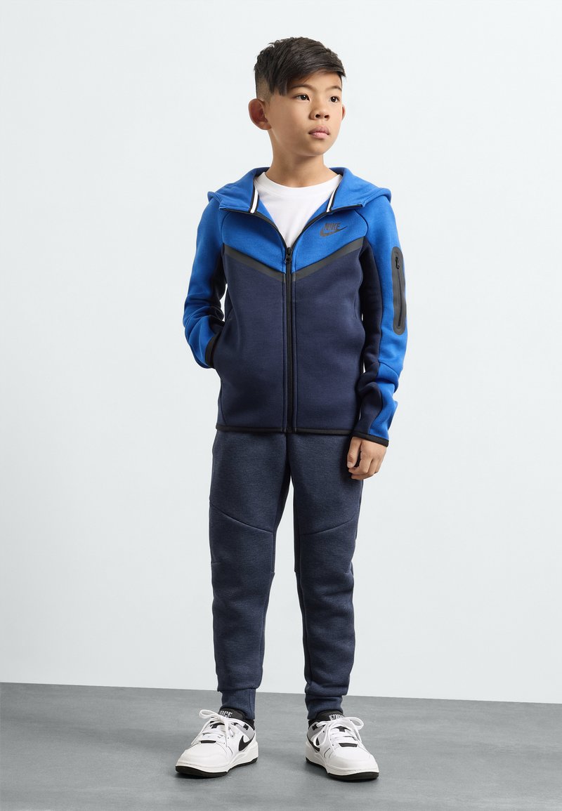 Blaue Zip-Hoodie mit grauen Akzenten, kombiniert mit navy Jogginghosen. Darunter ein weißes T-Shirt, abgerundet mit schwarz-weißen Sneakers.