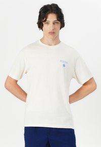 Crème-kleurig katoen t-shirt met korte mouwen en een ronde hals. Heeft een blauw grafisch logo op het linkerbovenborstgebied.