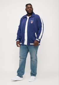 Blå varsityjakke med hvide striber, logo på brystet, trykknaplukning, sidelommer, parret med en hvid t-shirt og lyseblå jeans.