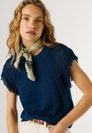 Mujer con cabello rubio ondulado que lleva una blusa de punto azul marino con mangas con flecos, pantalones beige, un cinturón marrón y un pañuelo verde con estampado en el cuello.