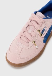 Sneakers Puma in morbido suede rosa pallido con accenti blu, caratterizzati da dettagli in pizzo, hardware dorato e un charm perla. Suola in gomma per una buona aderenza.