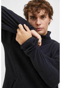 Pull en polaire noir avec col montant, manches longues et poches latérales zippées. Le tissu semble doux et texturé, avec une coupe décontractée.
