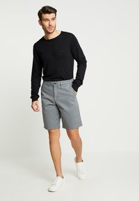 Harmaat shorts, joissa on nappikiinnitys ja vyölenkit, yhdistettynä mustaan pitkään hihaan ja valkoisiin tennareihin, seisoo neutraalilla taustalla.