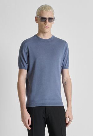 Jeune homme aux cheveux blonds courts portant des lunettes de soleil grises, un pull tricoté bleu à manches courtes et un pantalon noir sur un fond uni.