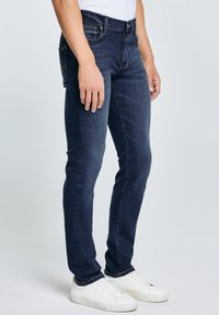 Osoba u uskim tamnoplavim trapericama i bijelim tenisicama, stojeći ispred jednobojne svijetlosive pozadine.